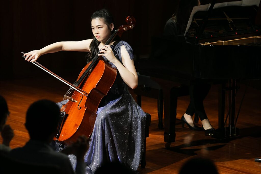 音樂會攝影:拉弓瞬間 穿藍紫色亮片禮服的大提琴演奏者拉弓,背景是鋼琴