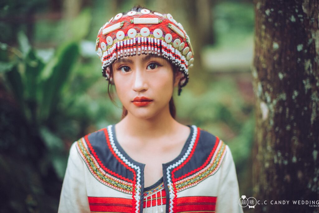 貝殼串珠頭飾 森林背景下的族服新娘特寫,戴紅色織布與白色貝殼圓片串珠頭飾,穿黑紅滾邊白色上衣