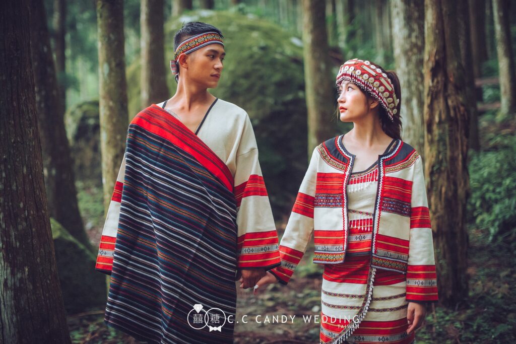 高大杉木林裡穿紅白條紋族服的新人牽手相望，新郎披深色條紋織布，新娘戴貝殼串珠頭飾