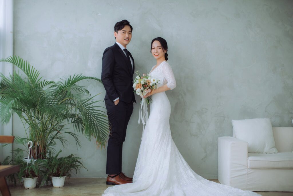竹北新人婚紗棚拍，蕾絲魚尾白紗新娘手捧花束與西裝新郎站在灰牆植栽旁