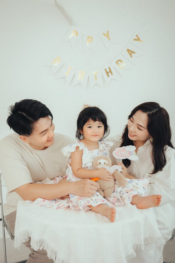 生日全家福|爸媽看著女兒 爸媽笑著看抱著樹懶玩偶的女兒,背景是 HAPPY BIRTHDAY 字母旗