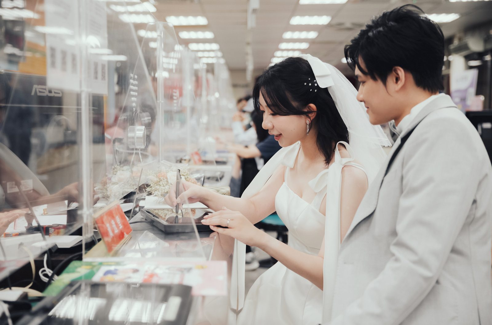 新娘在戶政櫃台簽名、新郎在旁微笑|結婚登記攝影