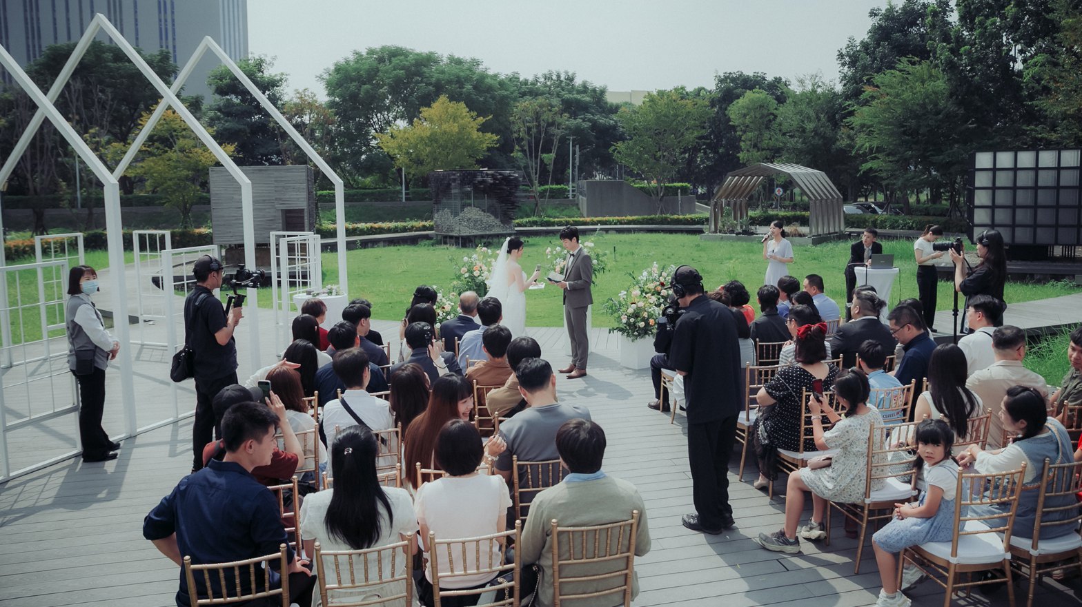 戶外證婚全景：新人在草地前宣讀誓詞，賓客圍坐、主持人拿麥克風站在一旁