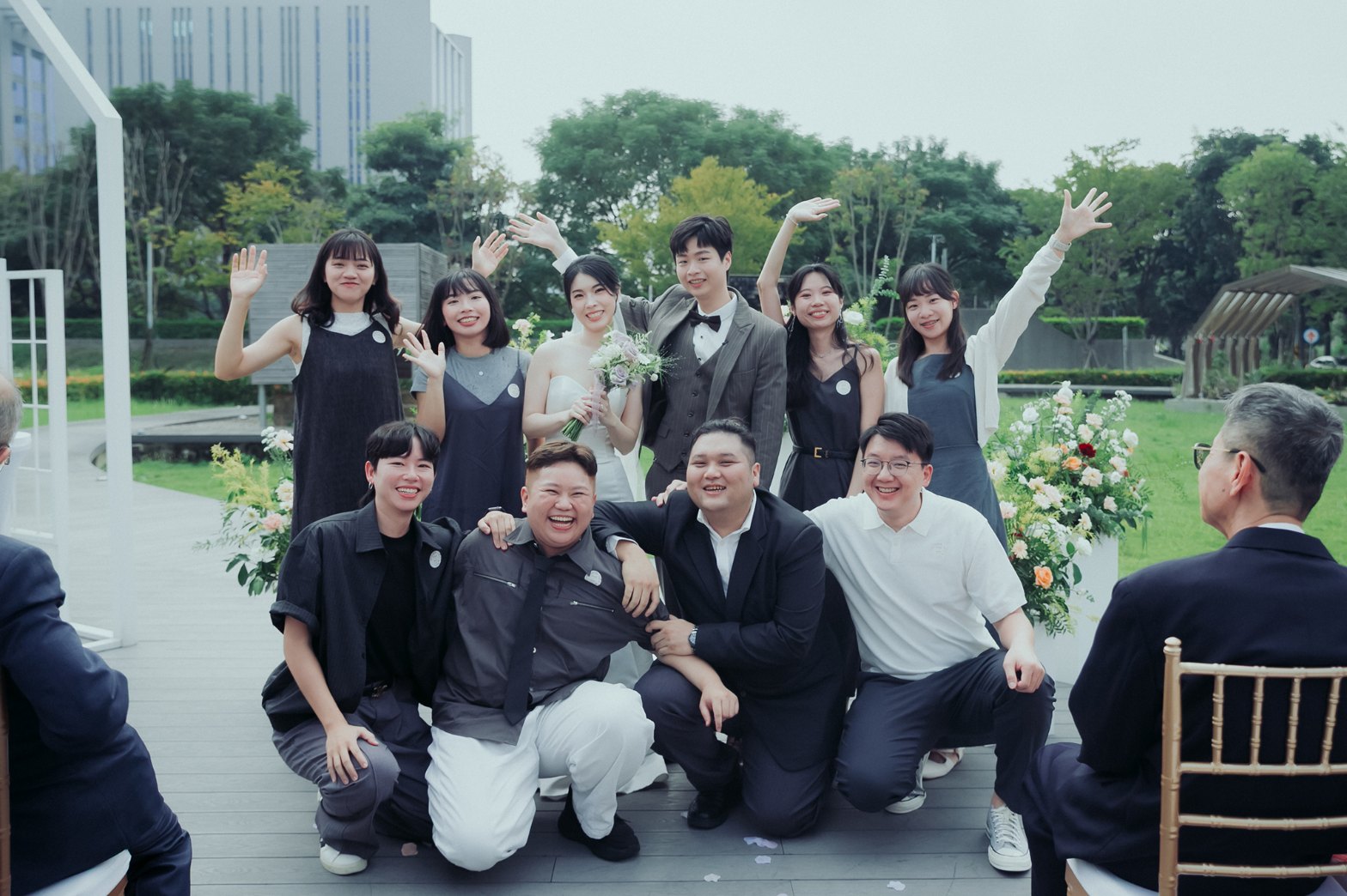 婚禮大合照：新人與朋友在草地前舉手揮手、笑得很開心