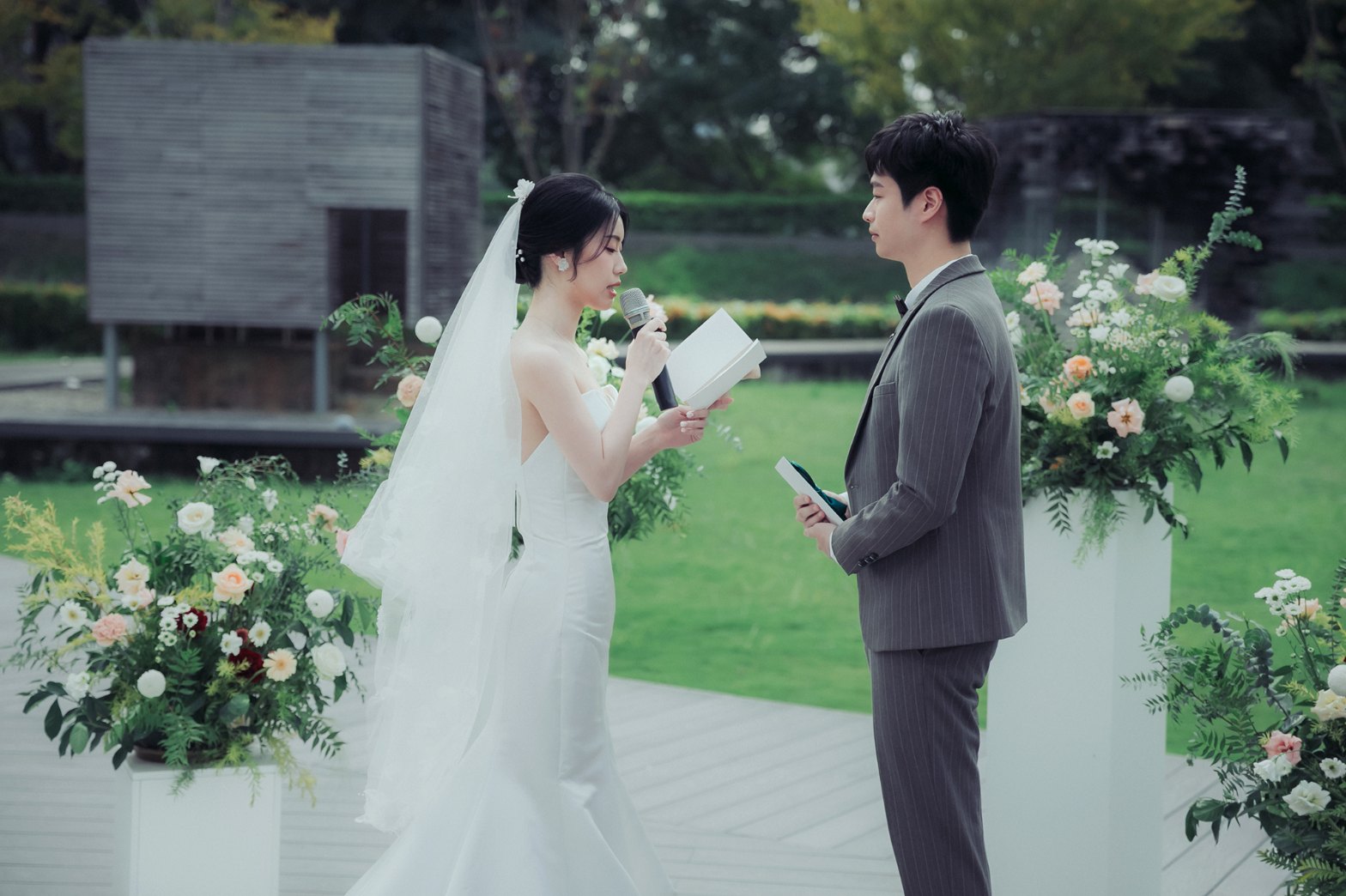 戶外證婚誓詞：新娘拿著麥克風唸誓詞，新郎在旁聆聽，以草地與花柱為背景