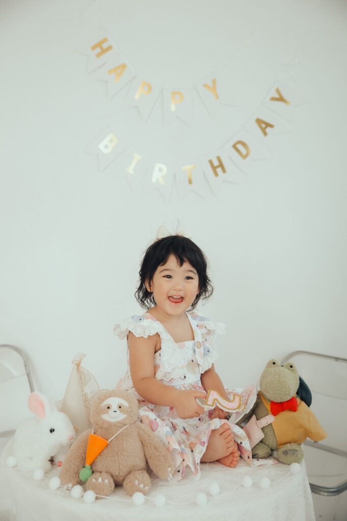 兩歲生日寫真|開心大笑 穿碎花洋裝的兩歲女孩在生日佈置前開心大笑,身旁是兔子和樹懶玩偶